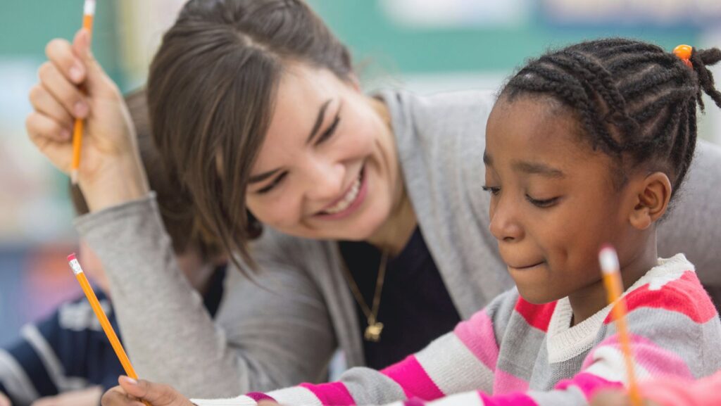 A teacher assists a young girl with her homework at a desk, fostering a supportive learning environment.